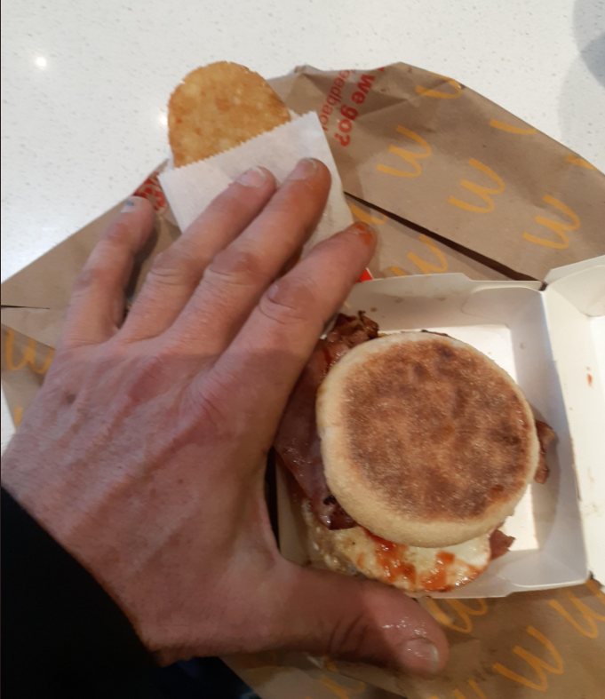 A hand holding a hash brown above an open container with an English muffin breakfast sandwich on a table with McDonald’s branded paper.