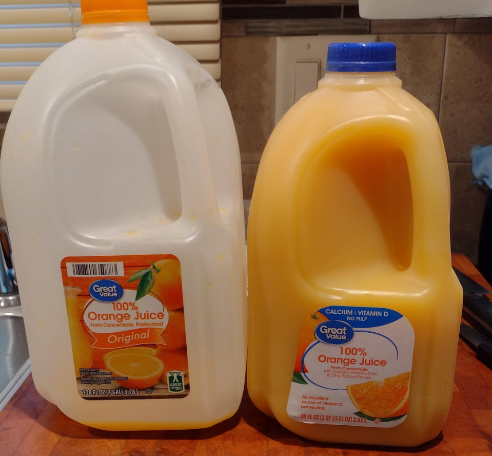 Two plastic jugs of Great Value orange juice are on a wooden counter. One is labeled "Original," the other "Calcium + Vitamin D." Both are partially empty. The background shows part of a sink and tiled wall.