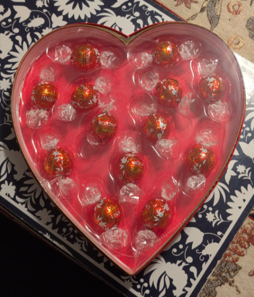 A heart-shaped box with red-wrapped Lindor chocolates arranged neatly inside, set on a dark and white patterned surface with part of a floral tablecloth visible in the corner.