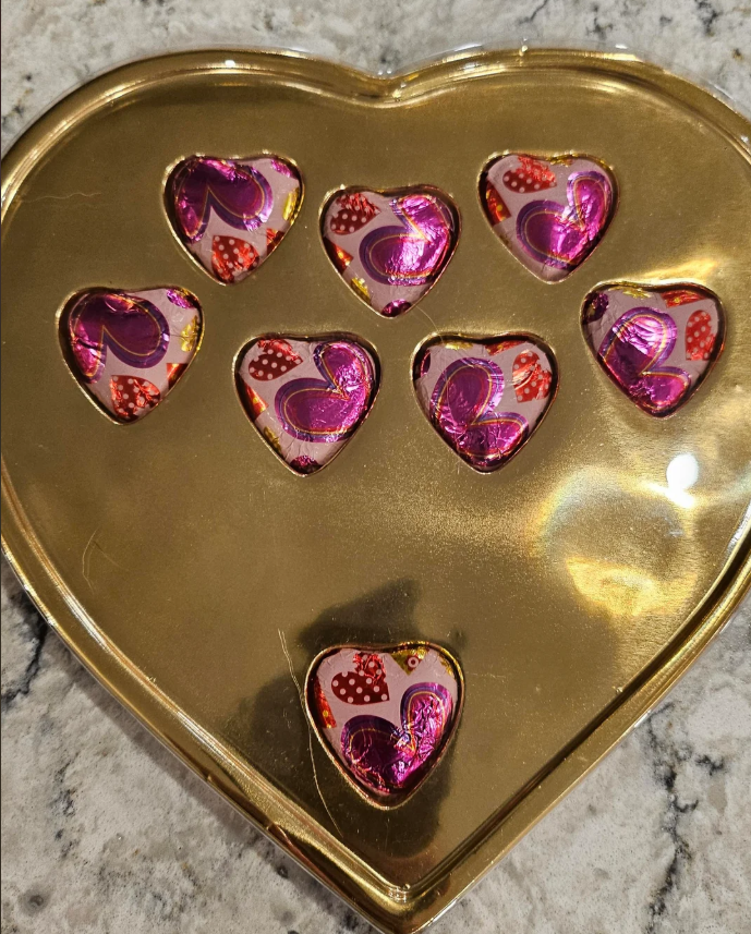 A gold, heart-shaped box with eight heart-shaped chocolates, each wrapped in shiny pink and red foil, sitting on a marble surface.