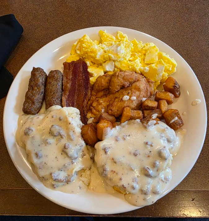 A hearty cheap all you can eat breakfast plate featuring scrambled eggs, chicken fried steak, sausage links, crispy bacon, home fries, and two biscuits smothered in sausage gravy on a brown tabletop.