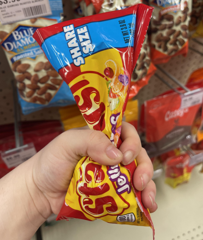 A hand is holding a share size bag of Starburst jellybeans in a store aisle, with other candy and snack bags visible on the shelves in the background.