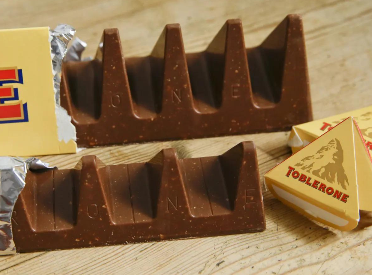 Opened Toblerone bars with their distinctive triangular chocolate segments are shown on a wooden surface, along with pieces of yellow packaging displaying the Toblerone logo and mountain design.
