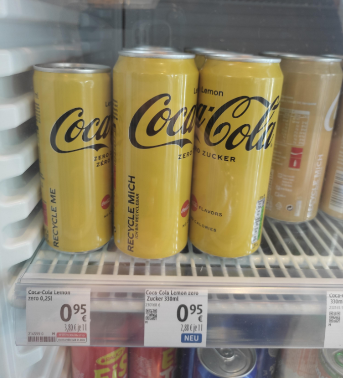 Three yellow cans of Coca-Cola Zero Sugar Lemon flavor are displayed in a refrigerator, with price tags below indicating 0.95 euros for a 330ml can. Other canned drinks are visible in the background.
