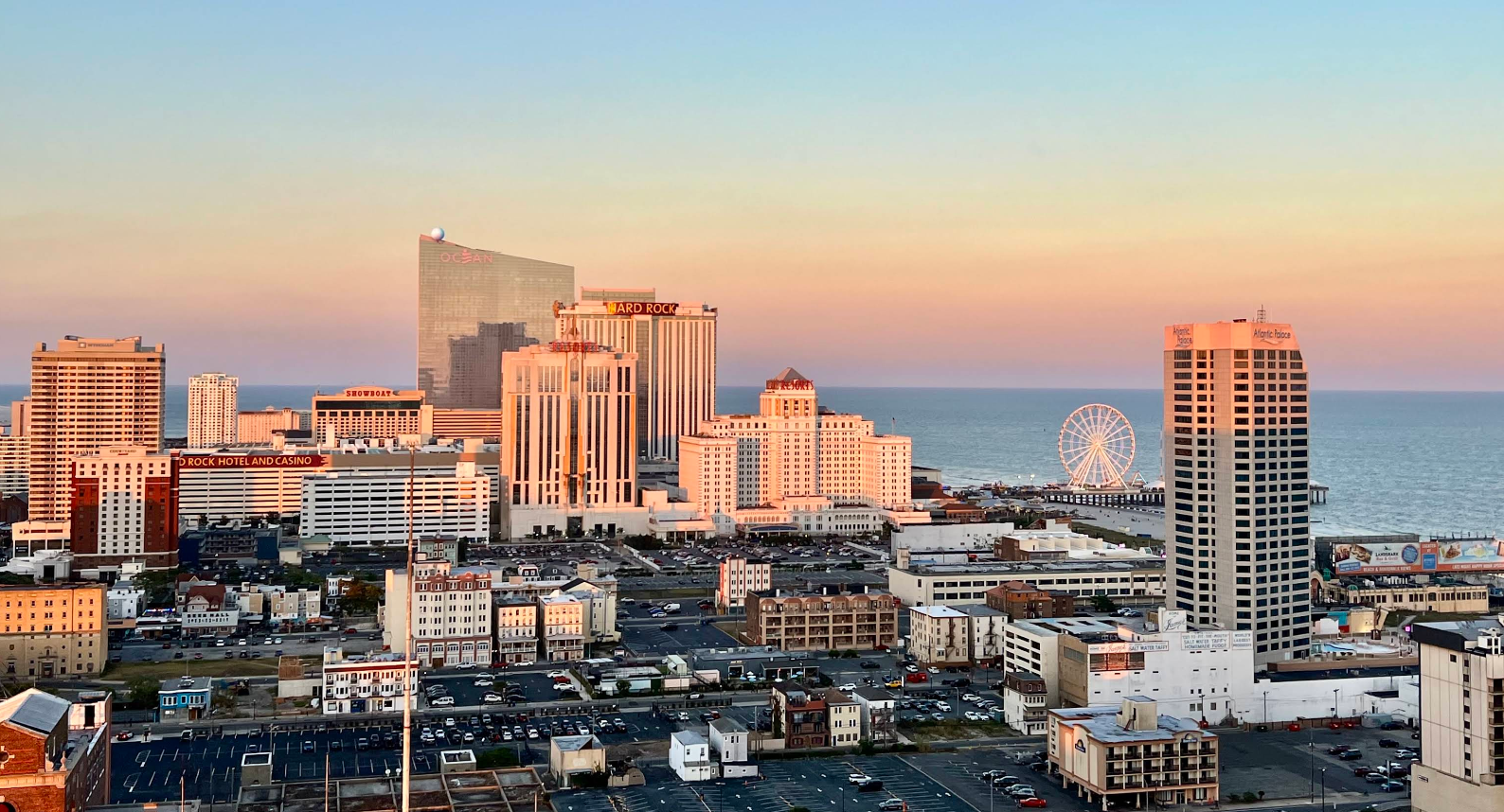 A cityscape at sunset with tall buildings, a ferris wheel near the ocean, and parking lots in the foreground. The sky is clear with a warm gradient from orange to blue.