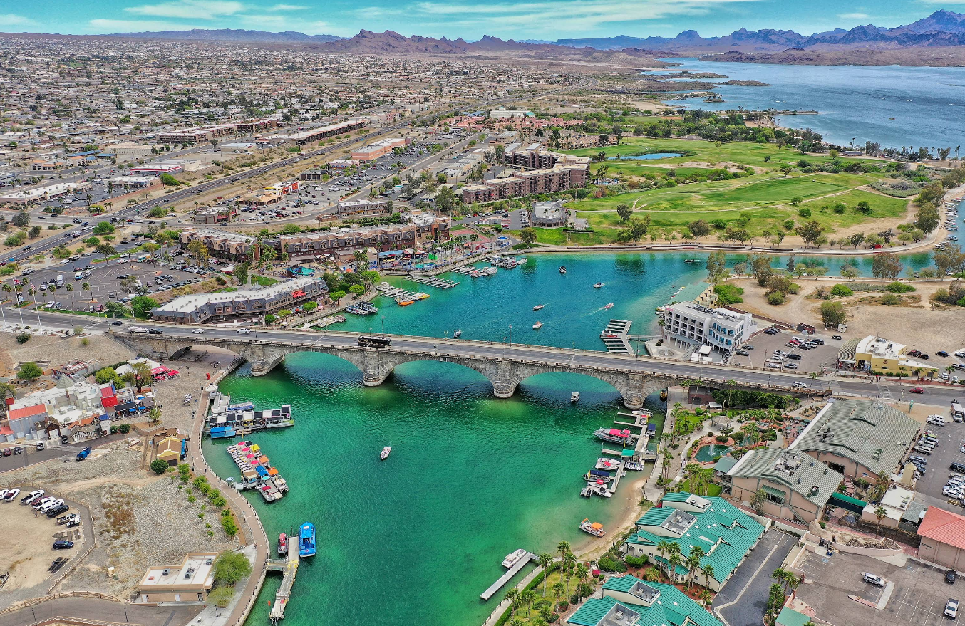 Aerial view of London Bridge crossing turquoise water in Lake Havasu City, Arizona, with boats, green parks, and surrounding cityscape visible under a blue sky.