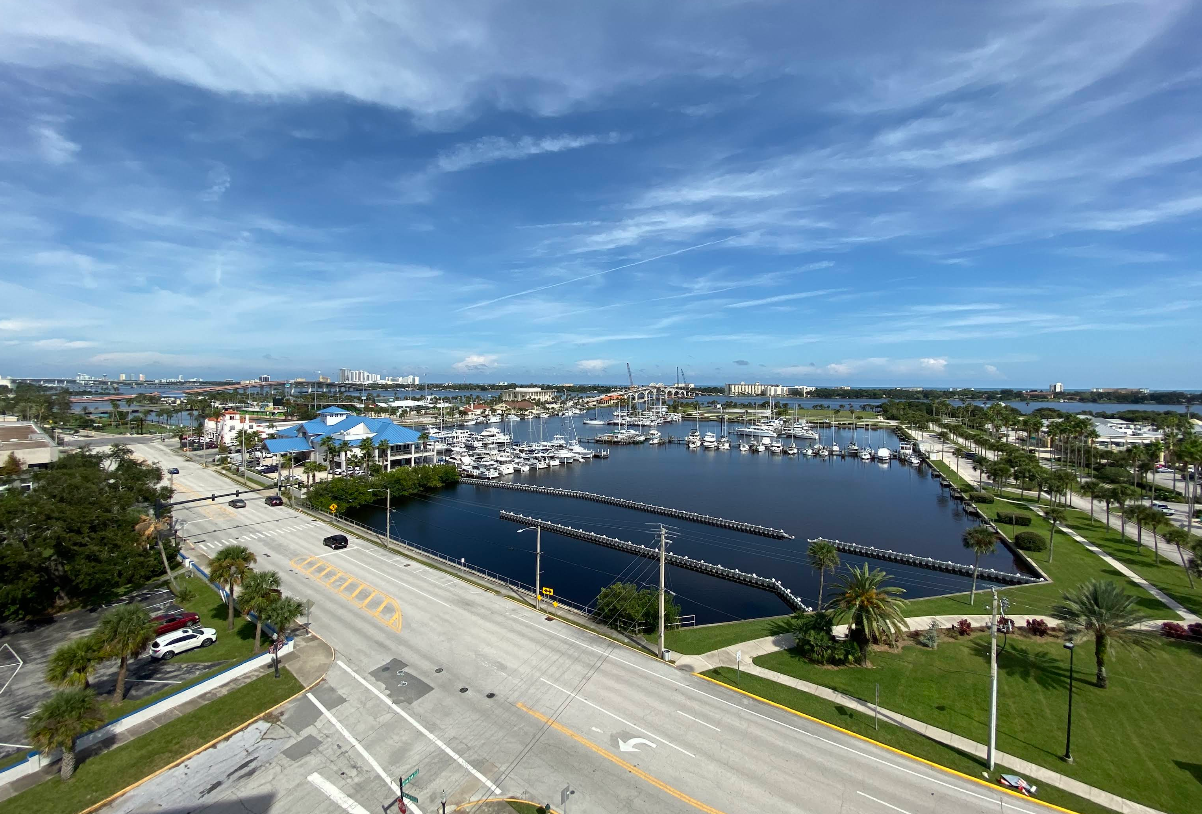A scenic view of a marina with many boats docked, surrounded by palm trees, roads, and buildings under a blue sky with scattered clouds.