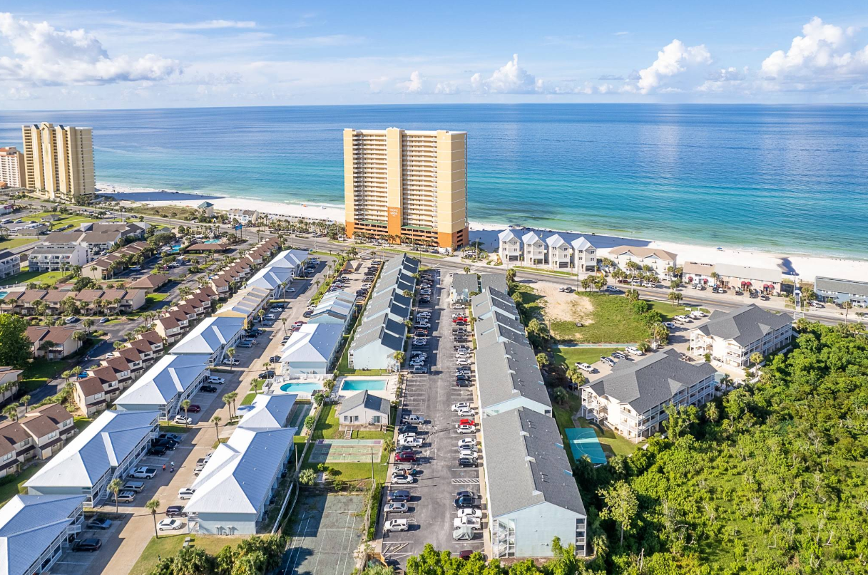 Aerial view of beachfront condos and apartments near a white sandy beach with turquoise ocean water, parked cars, a pool, and tall buildings under a clear blue sky.