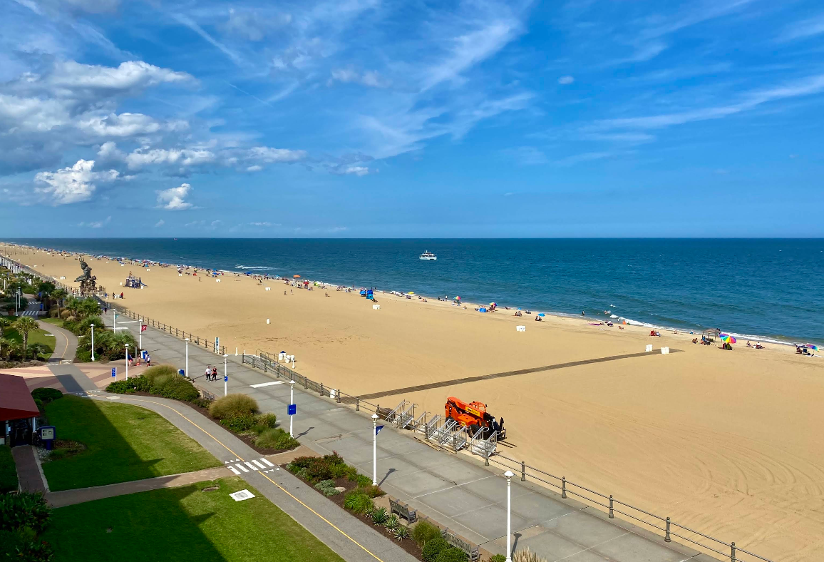 Wide sandy beach with a few people, a boardwalk, orange beach cleaning vehicle, green grass area, blue ocean, and a boat in the distance under a partly cloudy sky.