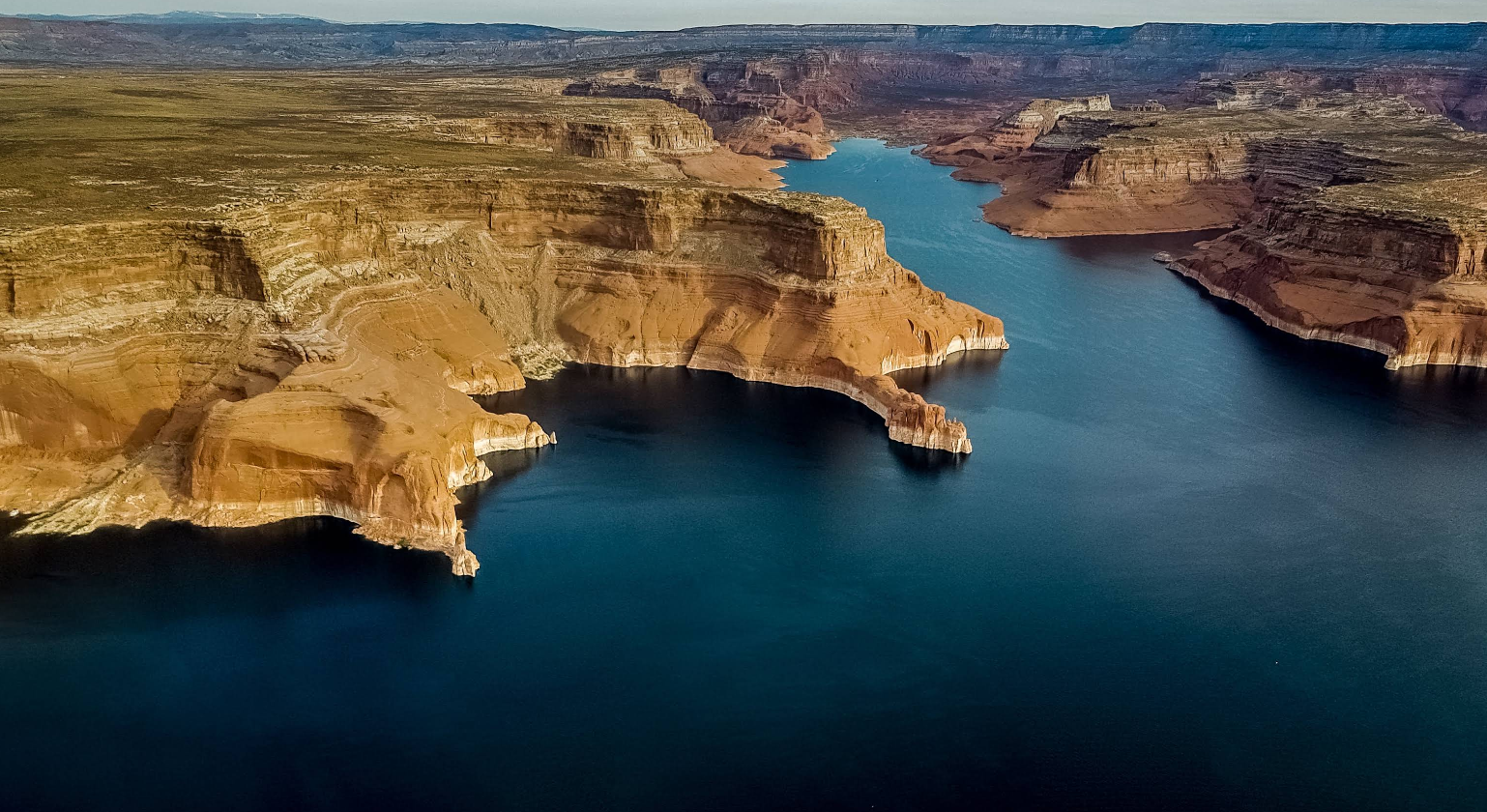 Aerial view of a winding river with steep, layered red rock cliffs and plateaus on both sides, under a clear sky.