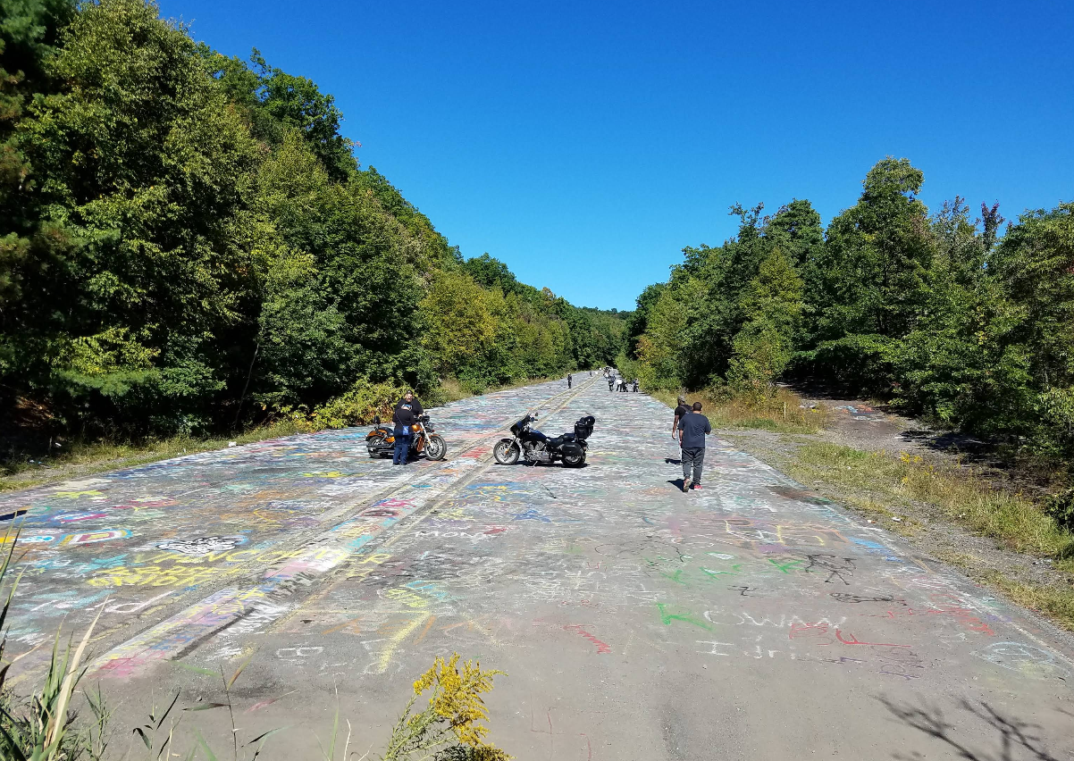 People stand by motorcycles on an abandoned, graffiti-covered road in the outskirts of one of those classic American towns, surrounded by green trees under a clear blue sky.