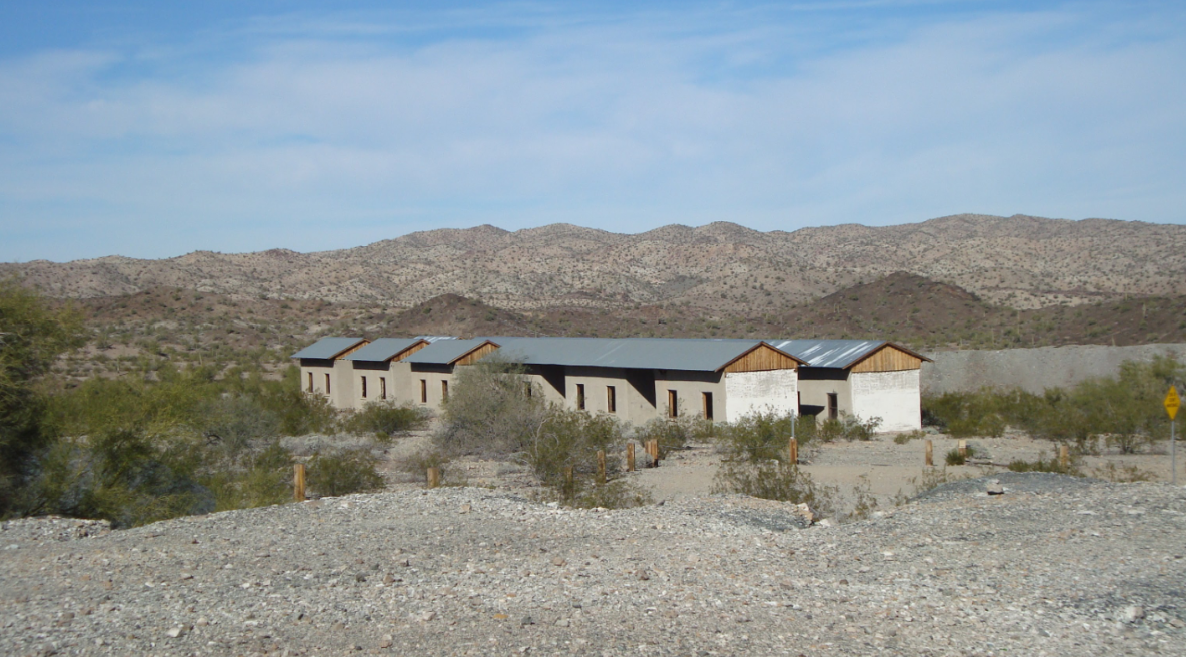 A long, abandoned building with a metal roof sits in a desert landscape—an evocative remnant often found in old American towns—surrounded by sparse bushes and rocky ground, with mountains in the background under a partly cloudy sky.
