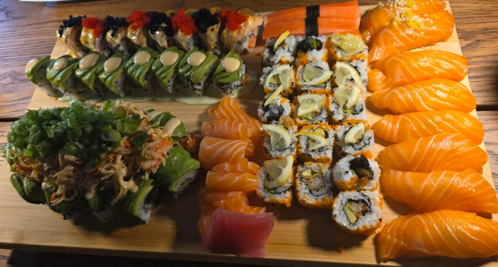 A wooden platter filled with assorted sushi rolls, nigiri, and sashimi, including salmon, avocado-topped rolls, crab meat, and pieces garnished with green onions and fish roe.