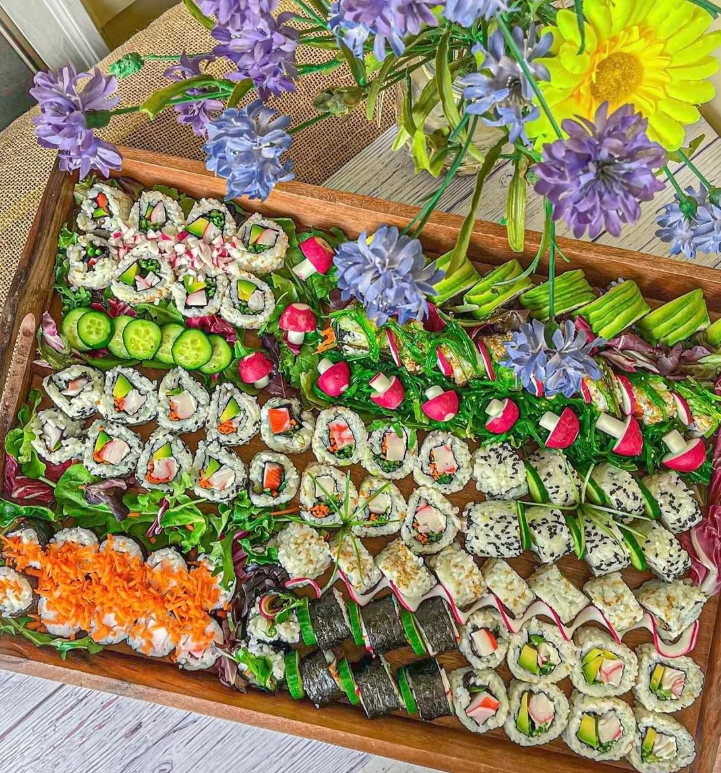 A wooden tray filled with assorted sushi rolls, garnished with vegetables and greens, sits on a table. Bright flowers in purple and yellow are arranged above the tray, adding color to the scene.