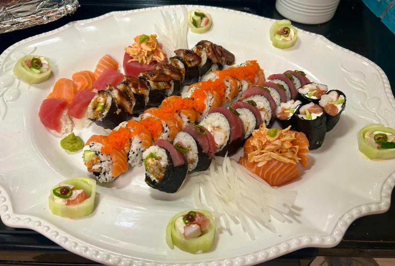 A white platter with assorted sushi rolls, sashimi, and garnishes, including sliced cucumbers filled with seafood, shredded radish, and small mounds of wasabi.