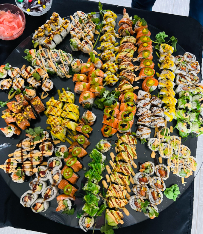 A large black platter filled with assorted sushi rolls, garnished with jalapeños, sauces, and greens, displayed on a black tablecloth. Pink pickled ginger is seen in a small bowl at the top left corner.