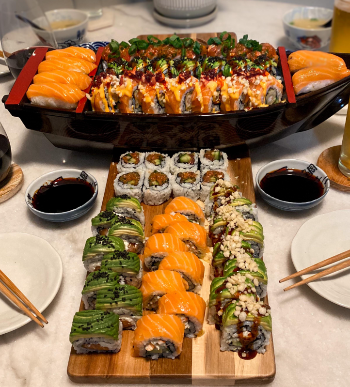 An assortment of sushi rolls topped with avocado, salmon, and sauces, served on wooden boards and a black sushi boat, surrounded by dipping sauces, drinks, and chopsticks on a table.