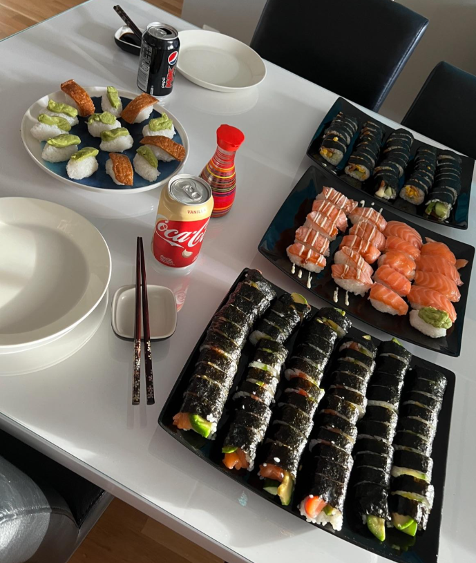 A table set with various sushi rolls, nigiri, a bottle of soy sauce, two cans of soda, chopsticks, and empty plates, ready for a meal. Four chairs surround the white table.