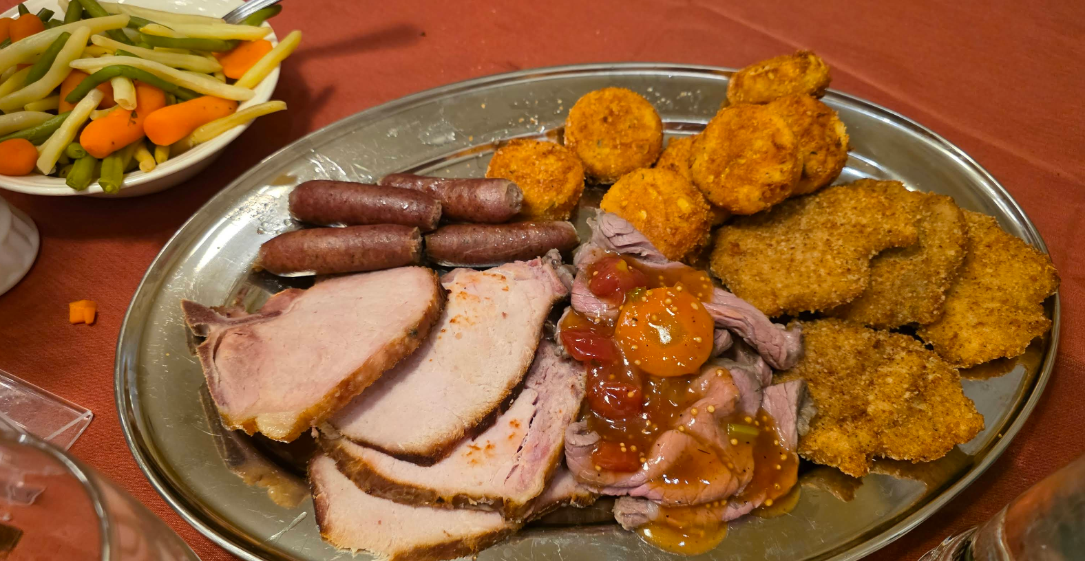 A silver platter with sliced roast pork, breaded cutlets, small sausages, and round breaded bites; a side bowl holds mixed pickled vegetables—all on a red tablecloth—perfect for a cheap all you can eat breakfast feast.