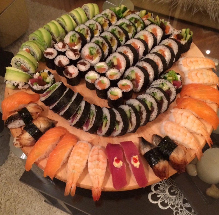 A round wooden platter with assorted sushi rolls, nigiri with salmon, shrimp, tuna, and eel, garnished with avocado and cucumber, sits on a glass table.
