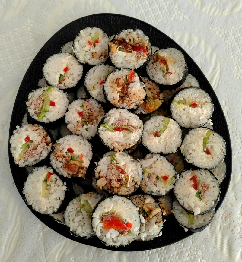 A black plate holds several pieces of sushi rolls filled with rice, vegetables, and possibly fish, arranged neatly on a white, textured tablecloth.