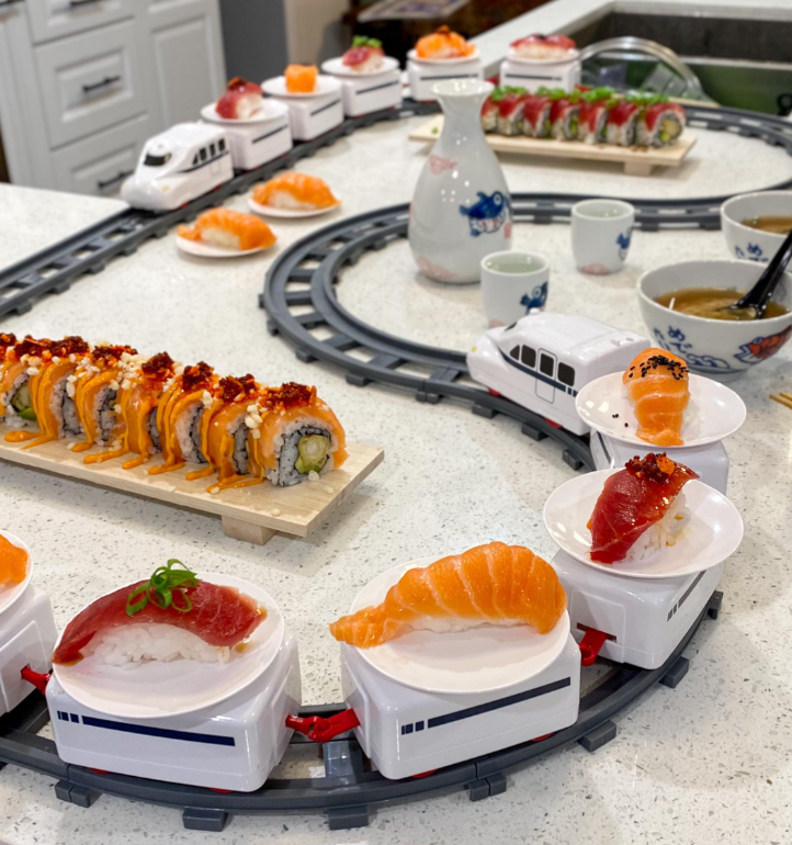 Sushi and sashimi plates are served on toy train cars circling a track on a kitchen counter, with bowls of soup, a sake bottle, and more sushi rolls in the background.