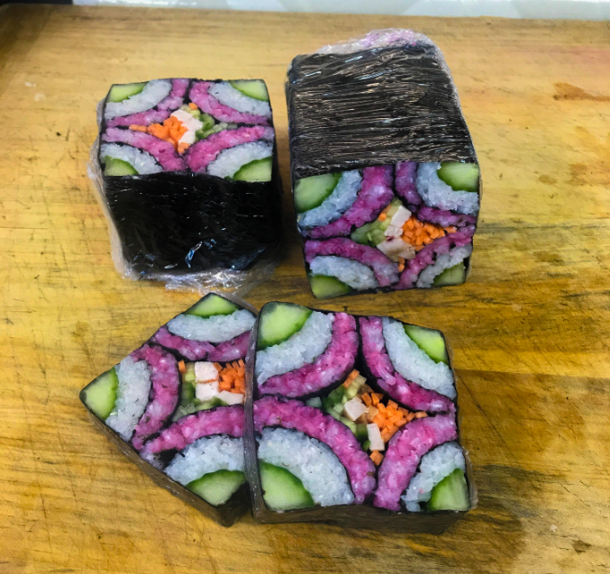 Three decorative sushi rolls with intricate, colorful patterns using rice, cucumber, carrot, pink and purple fillings, and nori sheets, shown on a wooden surface. Two pieces are sliced, revealing the patterned interior.