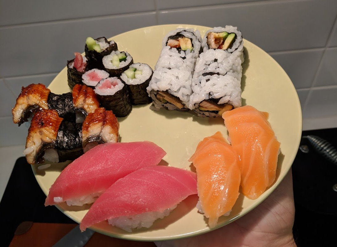 A plate of assorted sushi including nigiri with tuna and salmon, sushi rolls with cucumber and tuna, and rolls topped with cooked eel, against a tiled background.