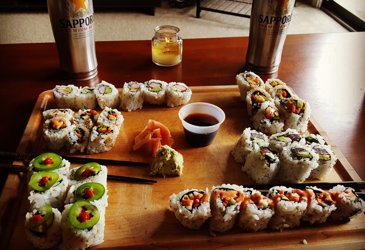 A wooden board with assorted sushi rolls, soy sauce, wasabi, pickled ginger, two Sapporo beer cans, and a lit candle on a table. Two pairs of chopsticks rest on the board.