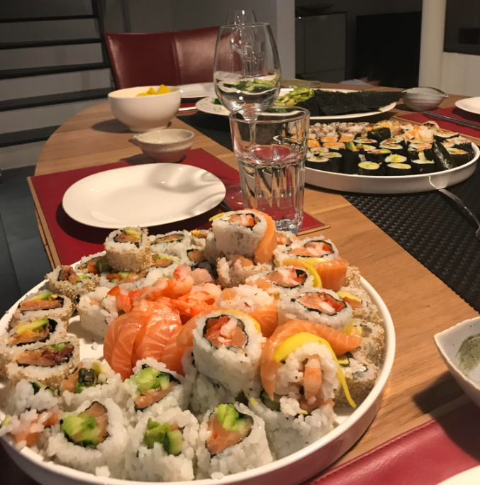A plate of assorted sushi rolls, including salmon and avocado, sits on a wooden table set with glasses of water, wine, and more sushi in the background.