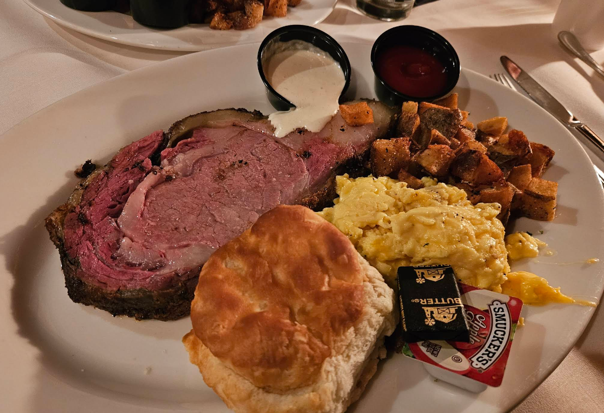 A plate from a cheap all you can eat breakfast features prime rib, creamy horseradish sauce, ketchup, scrambled eggs, breakfast potatoes, a biscuit, and packets of butter and jelly on a white tablecloth.