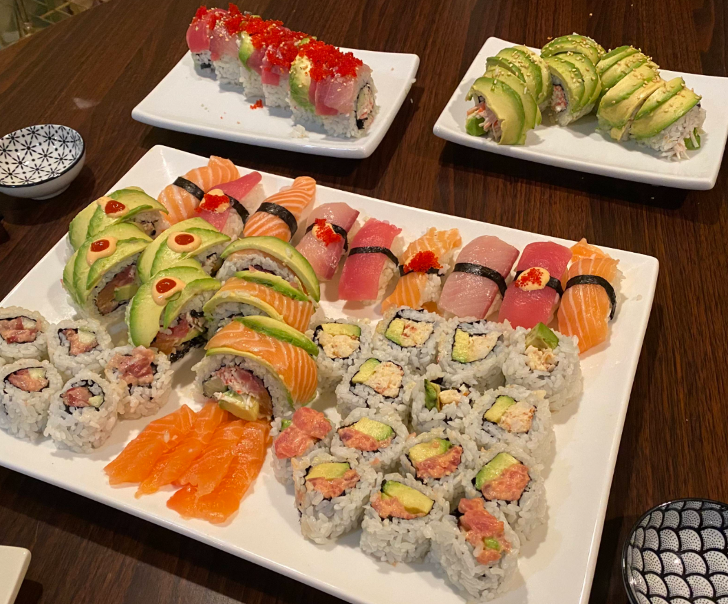 A large platter of assorted sushi rolls, sashimi, and nigiri is on a wooden table, with two smaller plates of specialty rolls and small dipping bowls. The sushi is colorful, featuring fish, avocado, rice, and garnishes.