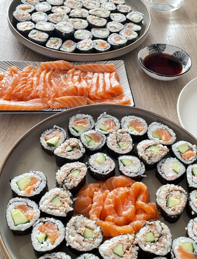 Three plates with sushi rolls and sliced salmon sashimi, a small dish of soy sauce, and a glass of water on a wooden table. The sushi features rice, cucumber, and salmon filling.