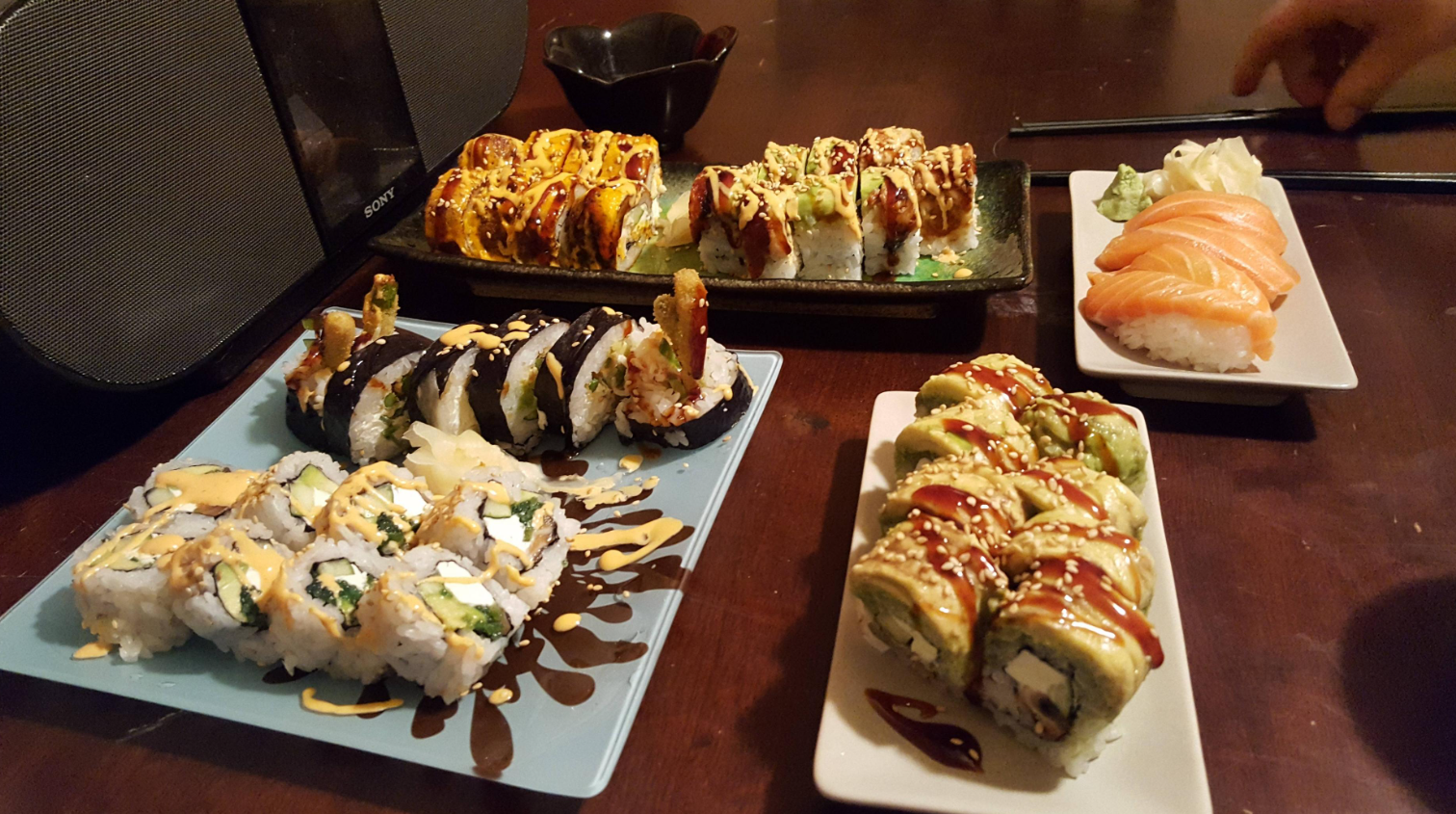 Four plates of assorted sushi rolls and nigiri sit on a dark wooden table, accompanied by wasabi and pickled ginger. A hand holds chopsticks in the background next to a large black speaker.