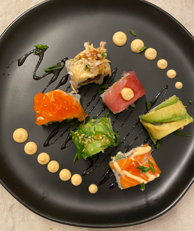 Six colorful sushi pieces topped with fish, avocado, and roe are artfully arranged on a black plate, accented with drizzles of sauce and dots of creamy dressing.