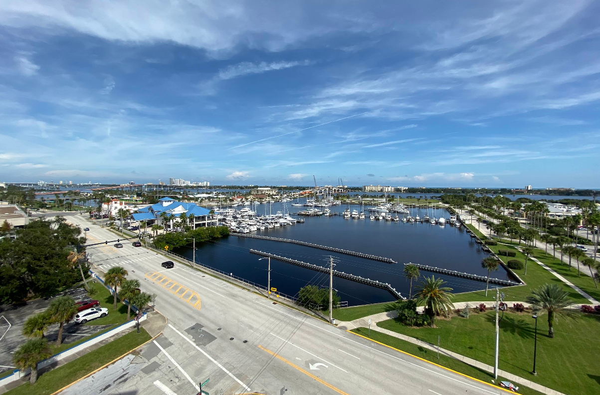 A marina with many docked boats, blue-roofed buildings, palm trees, green lawns, and a road in the foreground under a partly cloudy sky—this picturesque scene is one of the region's most inviting travel destinations.