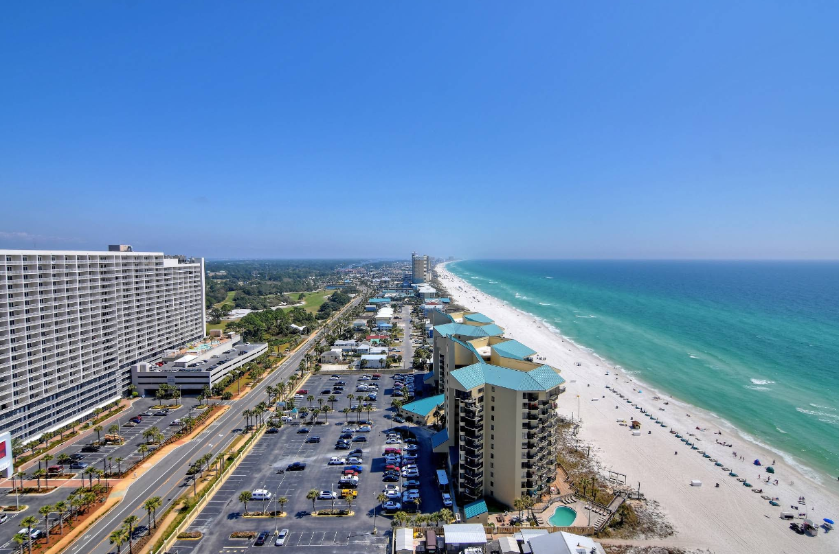 Aerial view of beachfront hotels, a parking lot, and a road beside a long, sandy beach with turquoise water under a clear blue sky—one of the most stunning travel destinations for those seeking sun and relaxation.