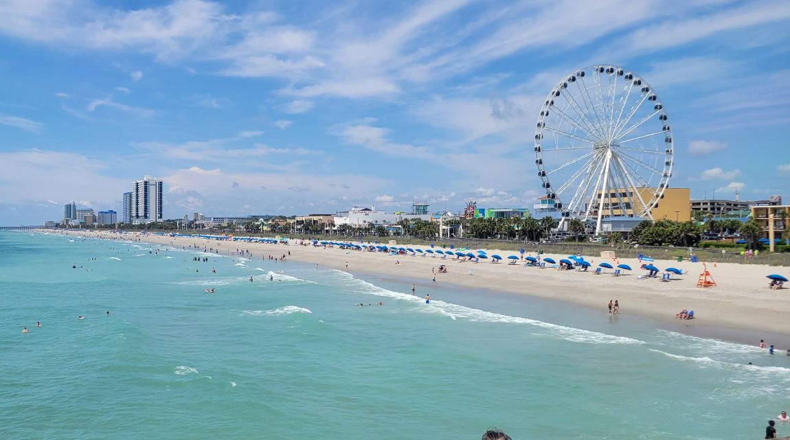 A sunny beach with people swimming and relaxing under blue umbrellas, a Ferris wheel, and tall buildings in the background under a blue sky with clouds—this vibrant scene perfectly captures one of the most inviting travel destinations.