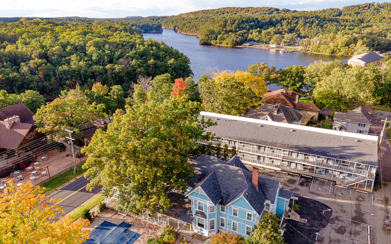 Aerial view of a blue Victorian house, nearby motel, and tree-lined street near a river—one of the region’s charming travel destinations—surrounded by dense green forest and hills under a clear sky.