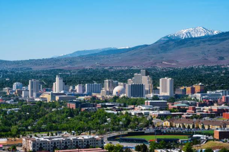 Cityscape of downtown Reno, Nevada, with modern buildings, green trees, and a stadium in the foreground. Snow-capped mountains are visible in the background under a clear blue sky, making it one of the most picturesque travel destinations for those seeking urban excitement against a stunning natural backdrop.