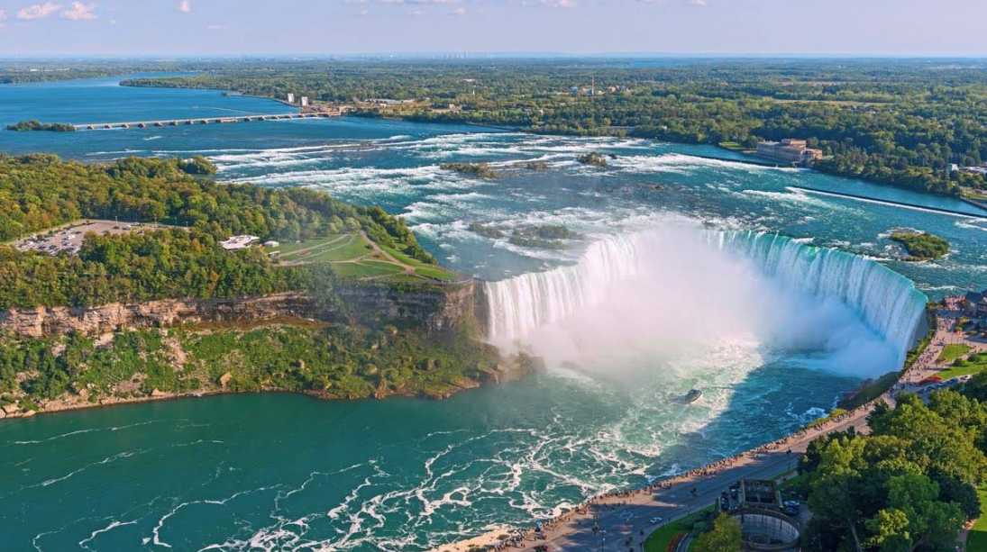 Aerial view of Niagara Falls with water cascading over cliffs, mist rising, lush green trees surrounding, and tourists gathered along the viewpoint—one of the world’s most breathtaking travel destinations.