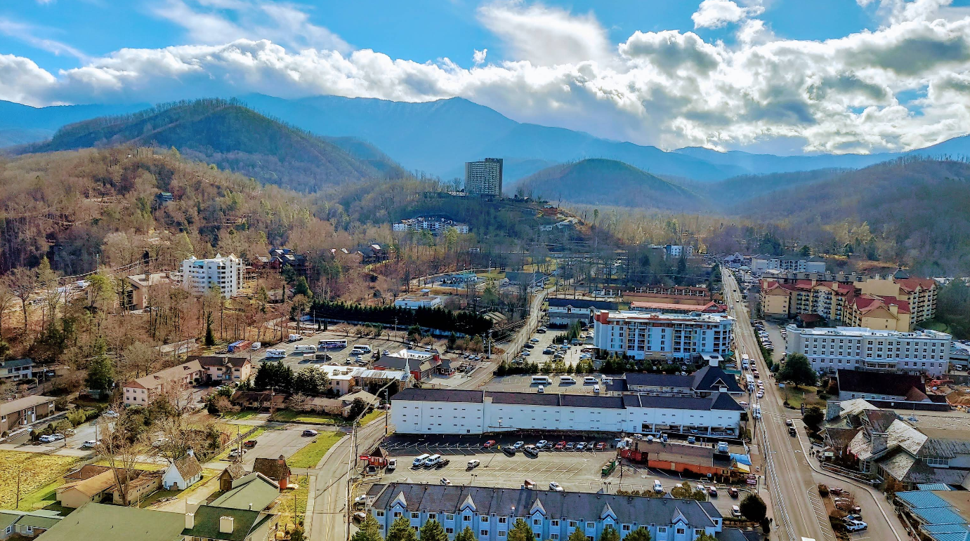 Aerial view of a small town with buildings, roads, and parking lots surrounded by forested hills and mountains under a partly cloudy sky—an inviting scene that highlights one of the region’s charming travel destinations.