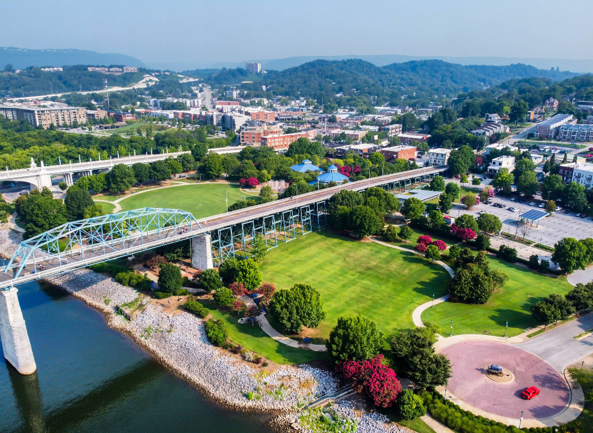 Aerial view of a city park, one of the top travel destinations, with a blue bridge, green lawns, trees, a roundabout with a red sculpture, buildings, and distant hills under a hazy sky.