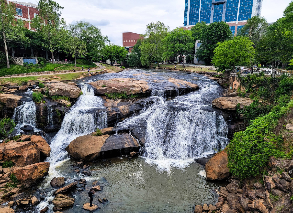 A multi-tiered waterfall cascades over rocks surrounded by greenery, with paths and modern buildings visible in the background on a cloudy day—making it a striking addition to any list of travel destinations.