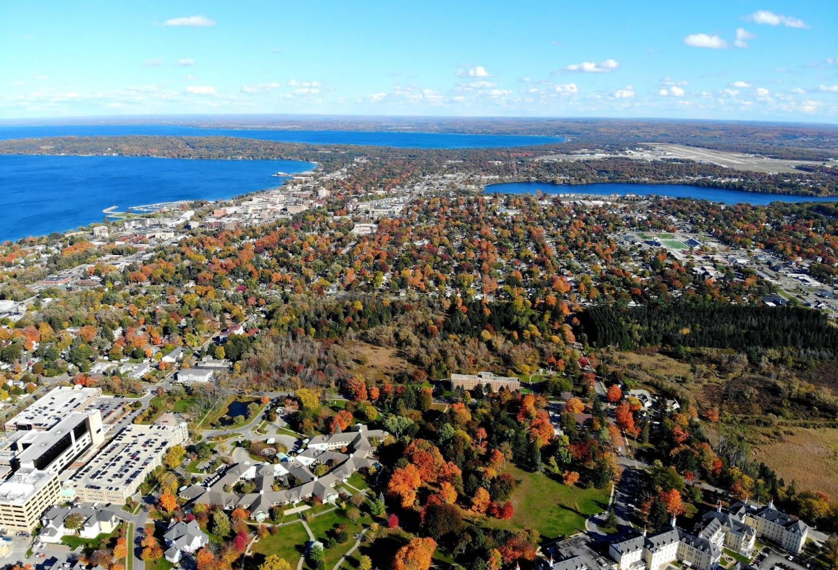 Aerial view of a town with colorful autumn trees, buildings, and parking lots, bordered by large blue lakes under a clear sky—one of the picturesque travel destinations perfect for a scenic getaway.