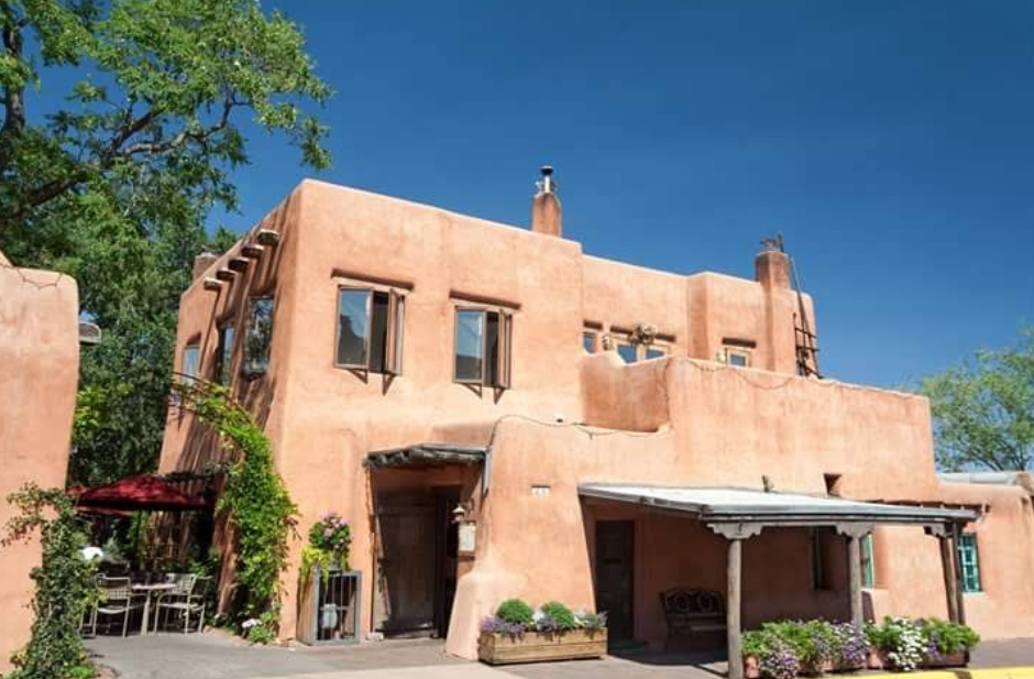 Adobe-style house with light brown stucco walls, wooden beams, and multiple windows, set against a clear blue sky. With its lush greenery, potted plants, and a charming outdoor seating area featuring a red umbrella, this picturesque setting embodies the inviting charm often found in unique travel destinations.