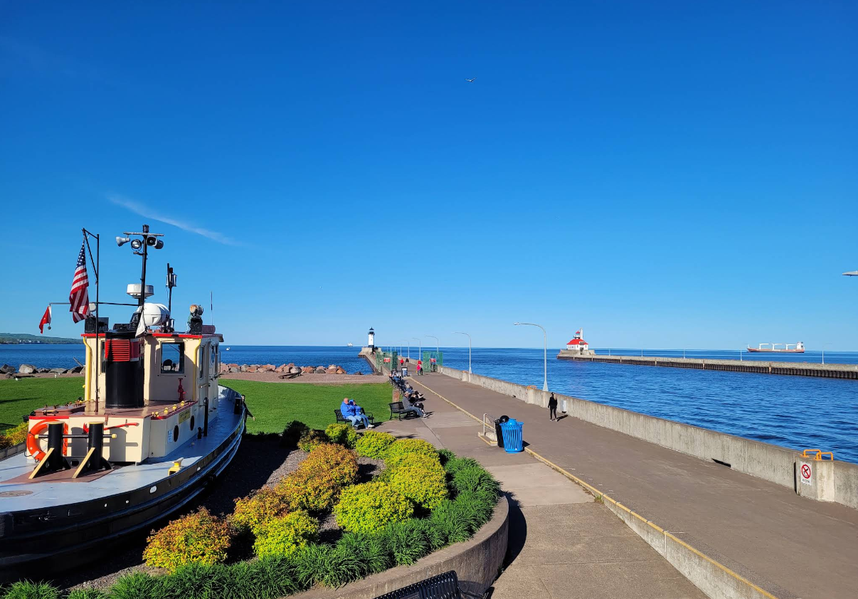 A small tugboat is docked by a walkway beside a blue lake, one of the charming travel destinations in the area. People stroll along the pier towards lighthouses. It’s sunny with clear skies, green grass, and neatly trimmed bushes in the foreground.