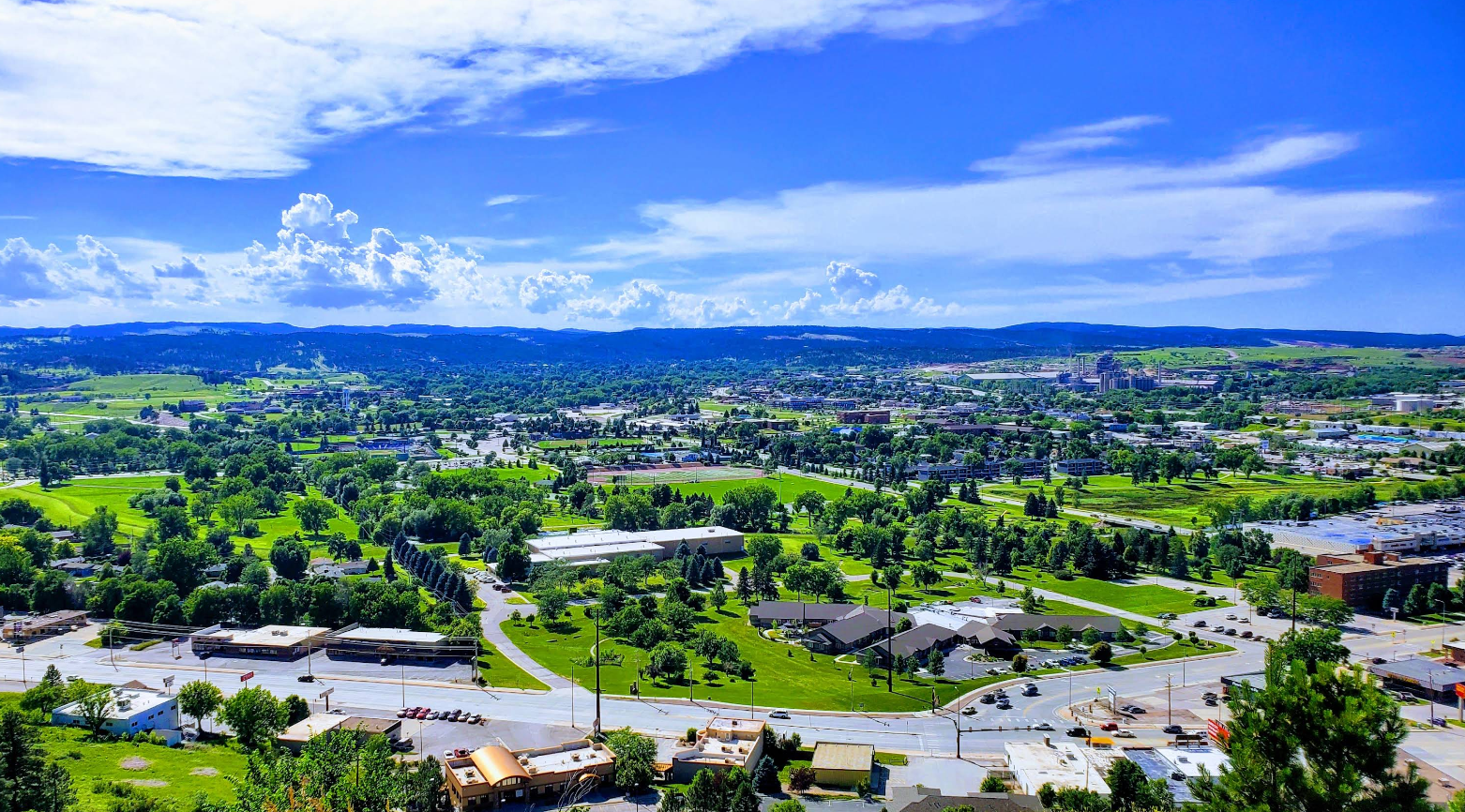 A vibrant cityscape with green trees, grassy areas, and buildings under a bright blue sky with white clouds, surrounded by distant hills—an inviting scene that captures the charm of one of the world’s most picturesque travel destinations.