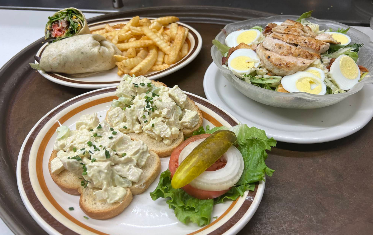 A tray with a chicken salad sandwich on toast with a pickle, a wrap with fries, and a green salad topped with grilled chicken, egg slices, and vegetables—perfect for enjoying after a cheap all you can eat breakfast.
