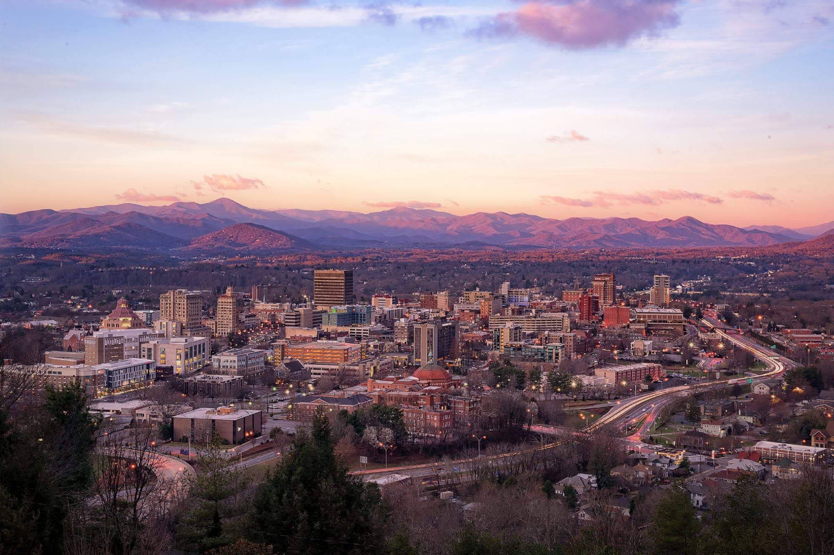 A cityscape at dusk with buildings, winding roads, and cars—one of the most captivating travel destinations. Mountains are visible in the background under a pink and purple sky with scattered clouds.